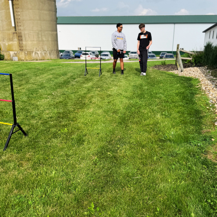 Two people standing on a grassy field playing ladder ball with a bridge and cars in the background