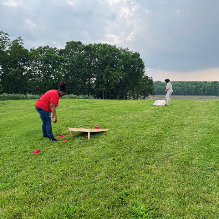 Two people playing cornhole on a grassy field with trees in the background