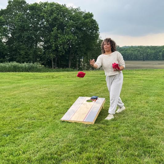 Woman playing cornhole in a grassy field with trees in the background