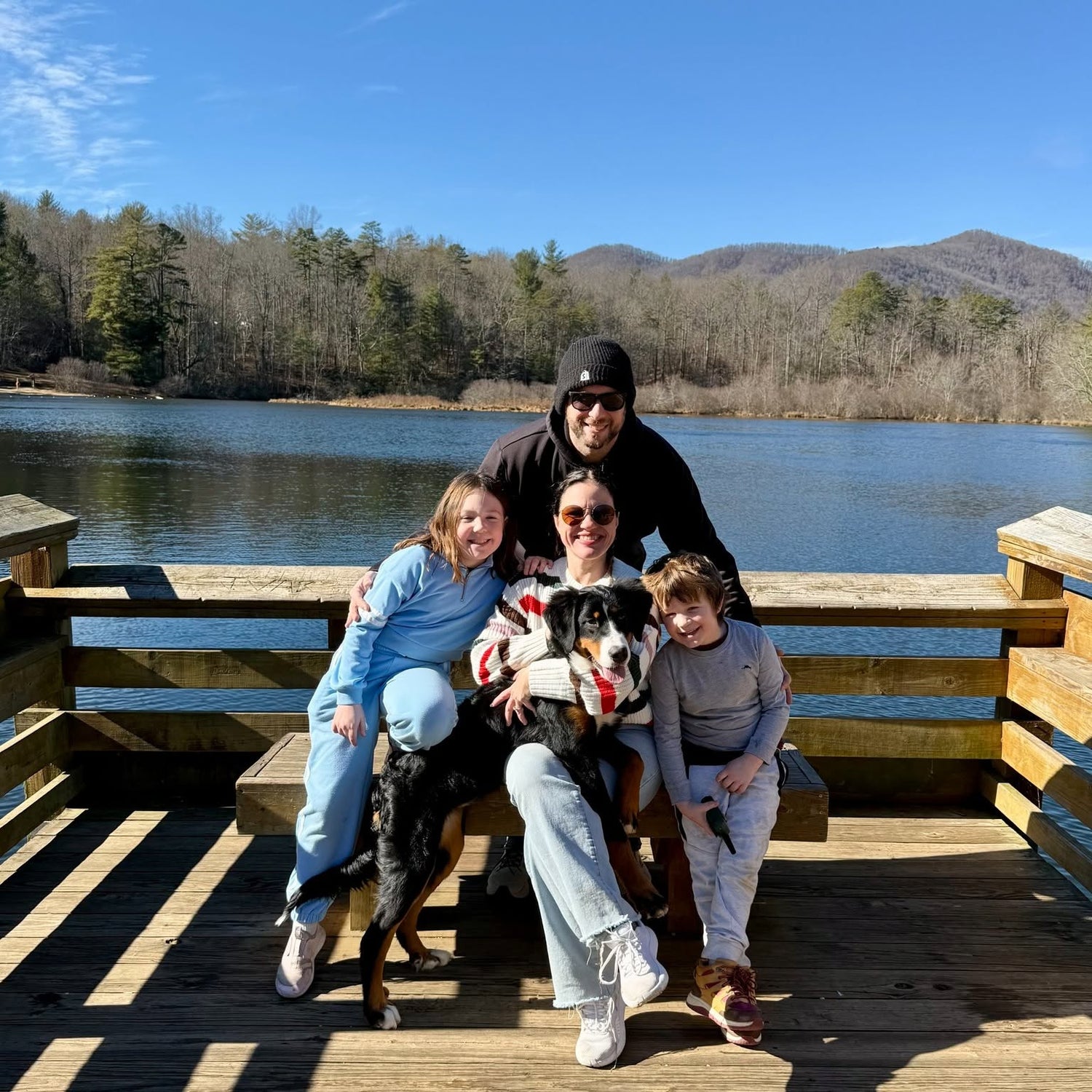 Family of four with a dog sitting on a wooden dock by a lake with mountains in the background.